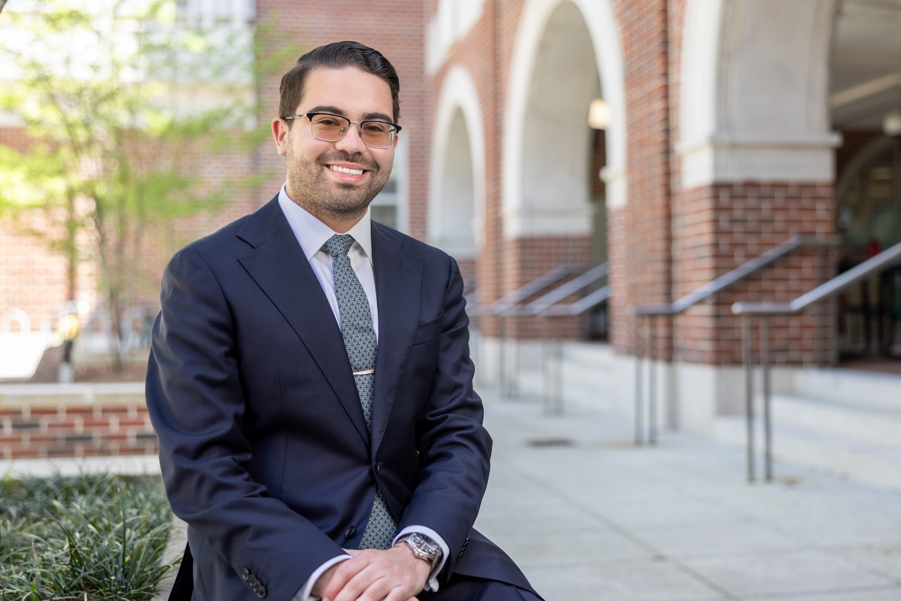 Smiling man in a dark suit and glasses sitting outdoors near a brick building.