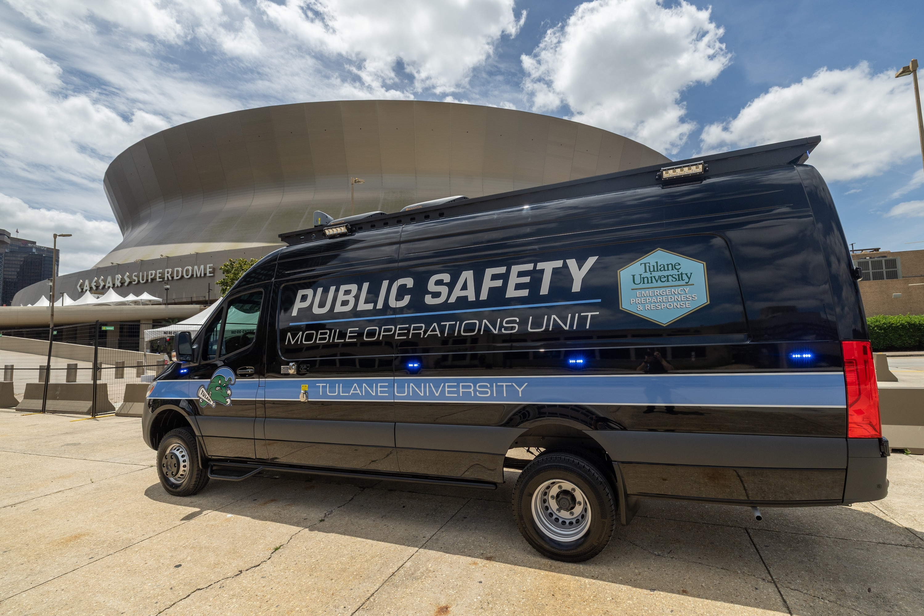 Black Tulane University Public Safety mobile unit van with blue lights, parked near a dome stadium.