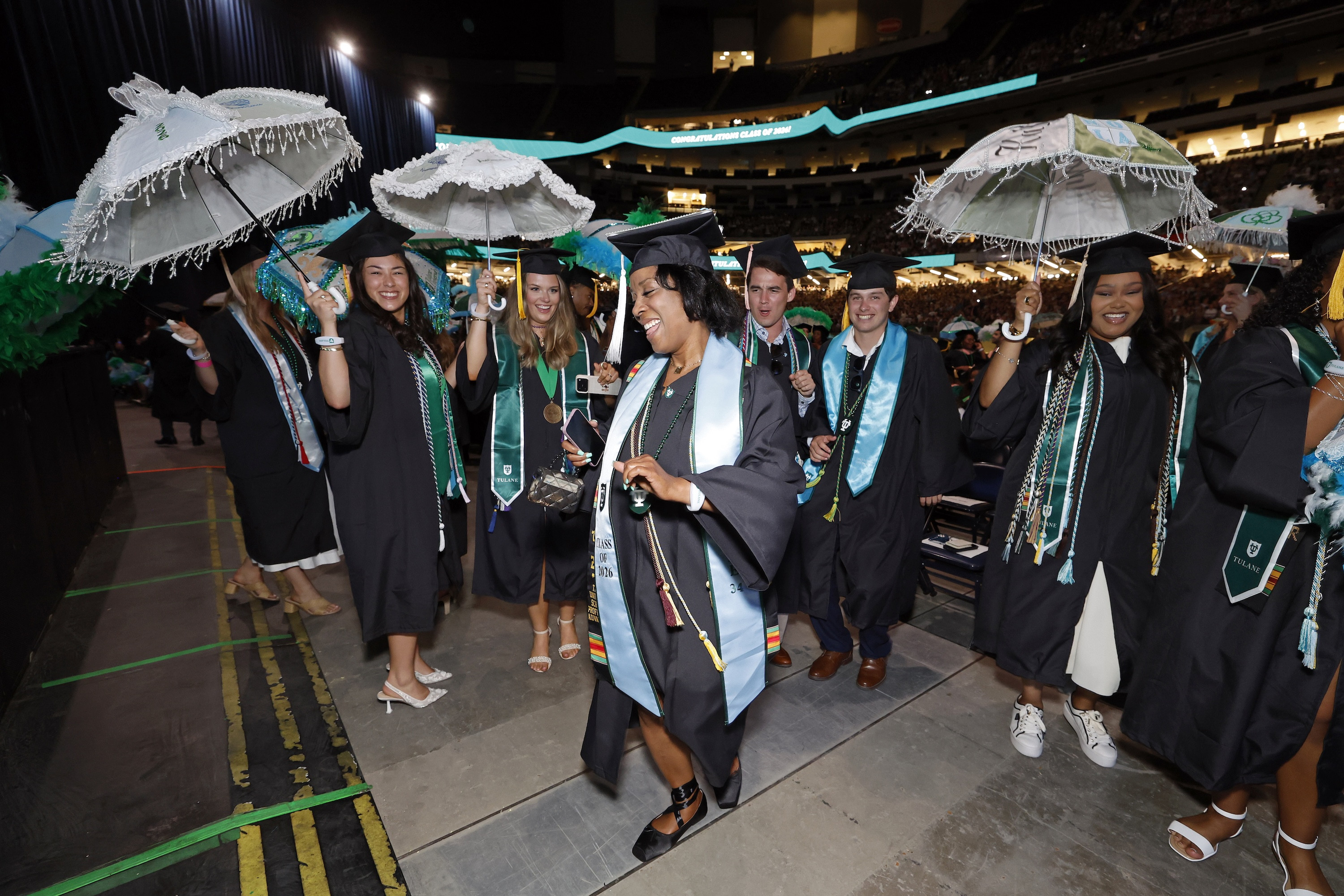 Joyful graduates in caps and gowns walk, some holding decorative white umbrellas.