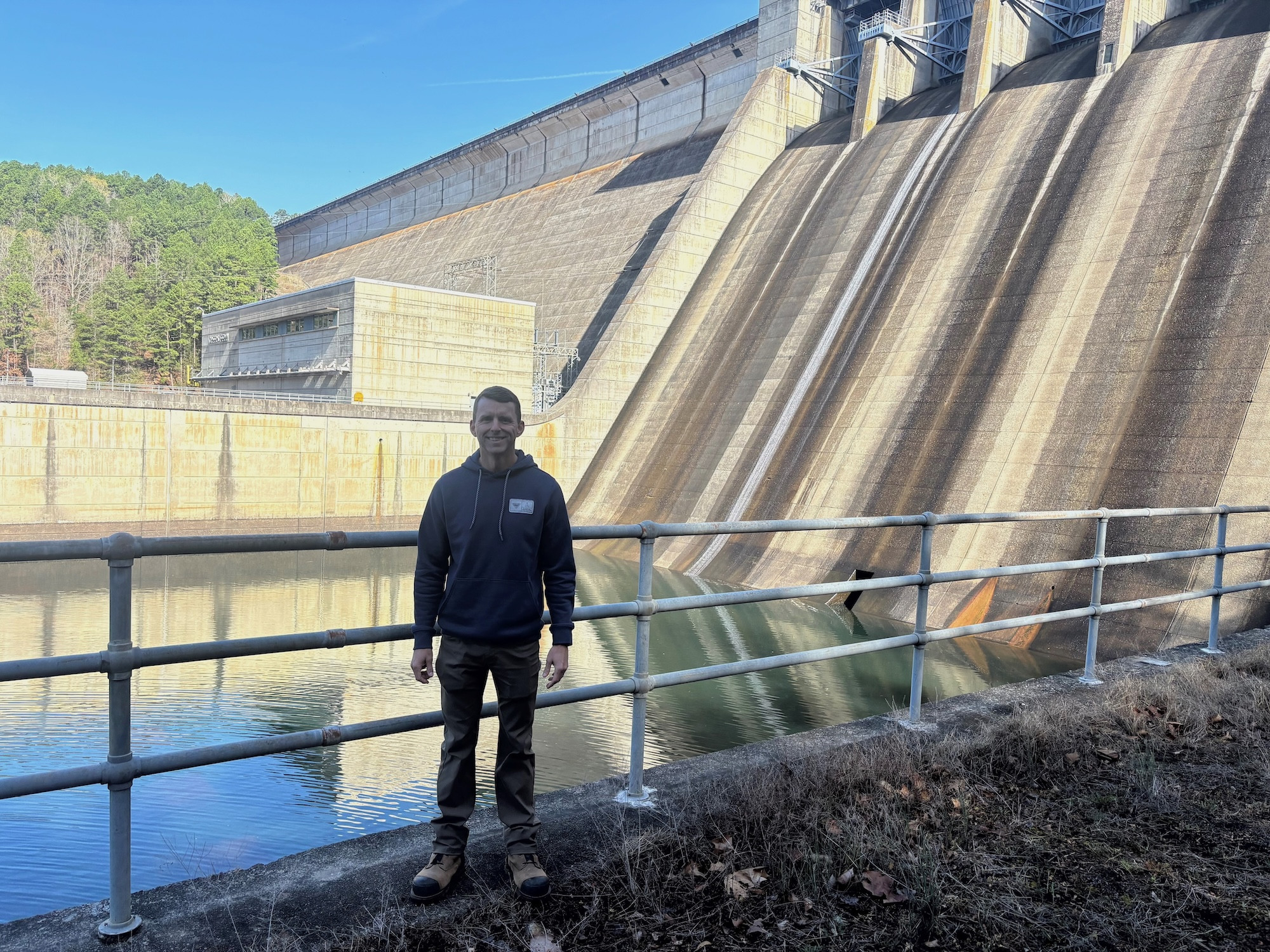 Man in a dark hoodie stands beside a massive concrete dam with water below on a clear day.