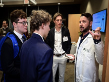 Bearded man in lab coat talks to three young men listening attentively at an event.