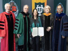 Four people in academic regalia and one woman in a suit, holding a document, pose by banners.
