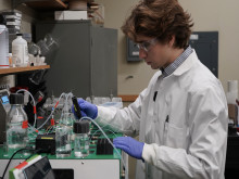 Young scientist in lab coat, glasses, and gloves, handling tubes on lab equipment.