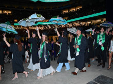 Joyful graduates in caps and gowns celebrating at night, holding umbrellas aloft in a stadium.
