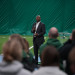 David Harris in dark suit speaks into microphone to audience on an indoor green field.