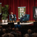 Two people sit on a stage with "BOOKFEST" banners and red curtains, speaking to an audience.