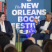 Two men on stage, one smiling, the other gesturing, with a New Orleans Book Festival backdrop.