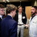 Bearded man in lab coat talks to three young men listening attentively at an event.