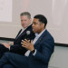 Gary Carter Jr. in blue suit speaking into a microphone, seated beside Patrick Connick listening.