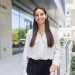 Smiling young woman in white blouse stands outside modern building.